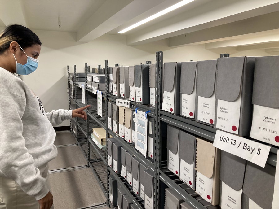 A young woman with dark hair and bleach streaks wearing a medical mask and light gray sweatshirt points and looks at archival boxes on shelves.