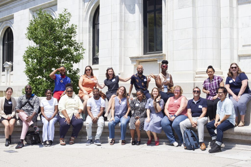 A group of 18 male and female teachers pose smiling and sitting outside a white marble building on a sunny summer day.