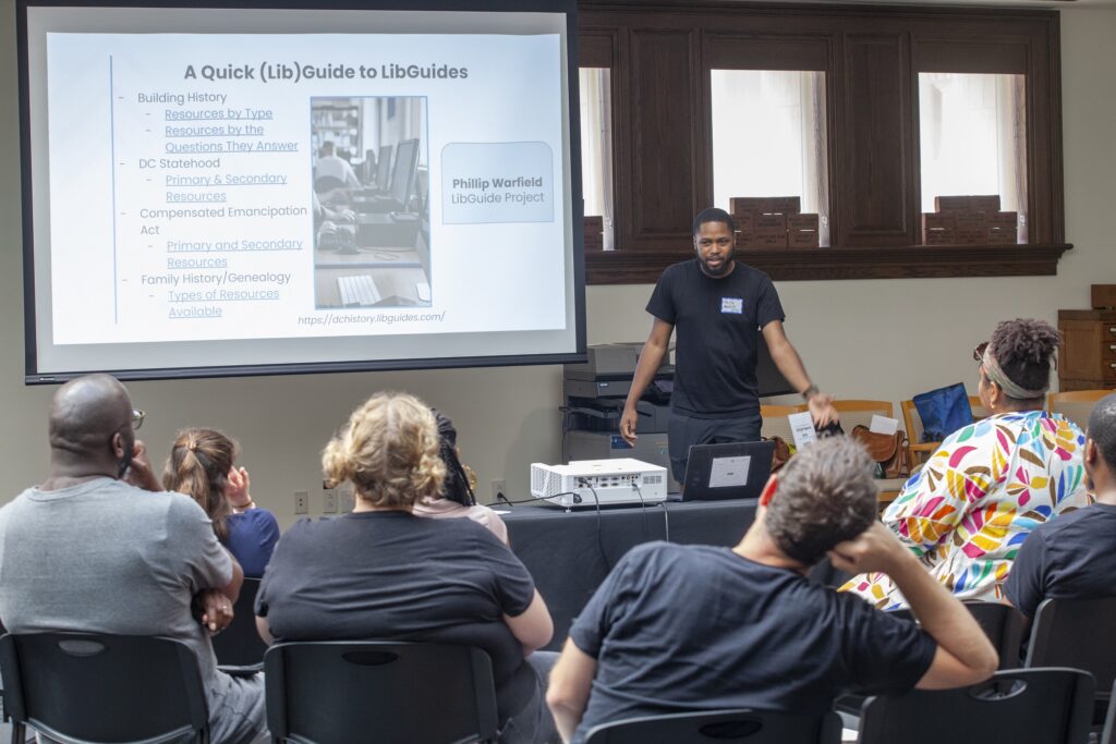 A man with a dark beard and short hair wearing a dark-colored t-shirt addresses a group of seated adults during a presentation. He stands next to a projector screen that reads "A Quick (Lib)Guide to LibGuides."