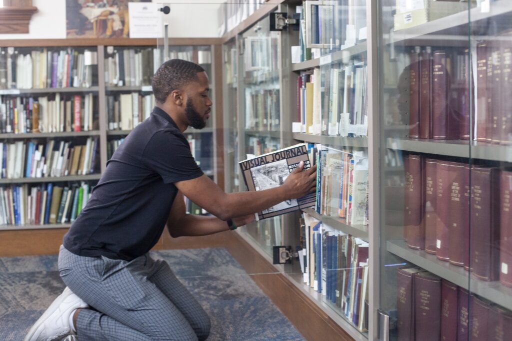 A man with a dark beard and short hair wears a dark blue t-shirt and grey checkered pants. He kneels while reshelving a book in a special collection's library,
