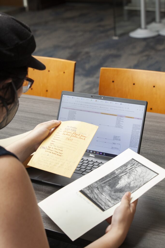 A person wearing cap and glasses viewed from behind sits at a table holding archival documents. A laptop is open in front of them.