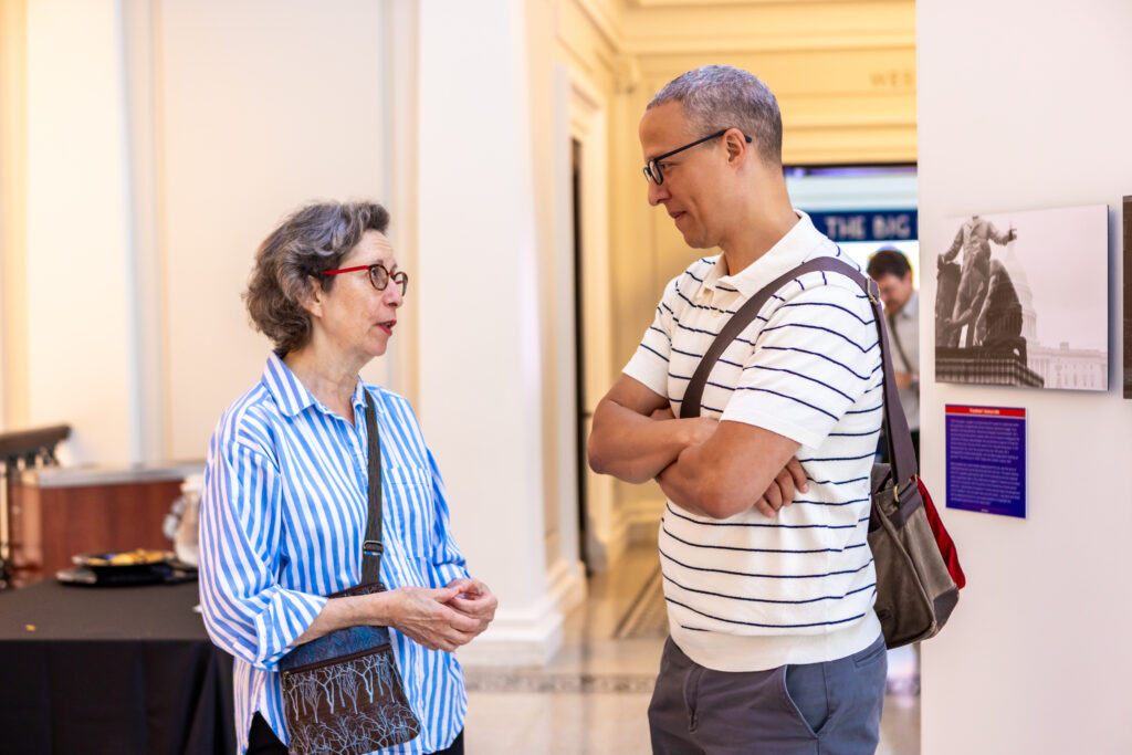 A woman wearing glasses, a blue and white vertical striped shirt, and cross-body purse stands in conversation with a man wearing a glasses, white polo shirt with blue horizontal stripes, and a messenger bag in the DC History Center's Memorial Hall.