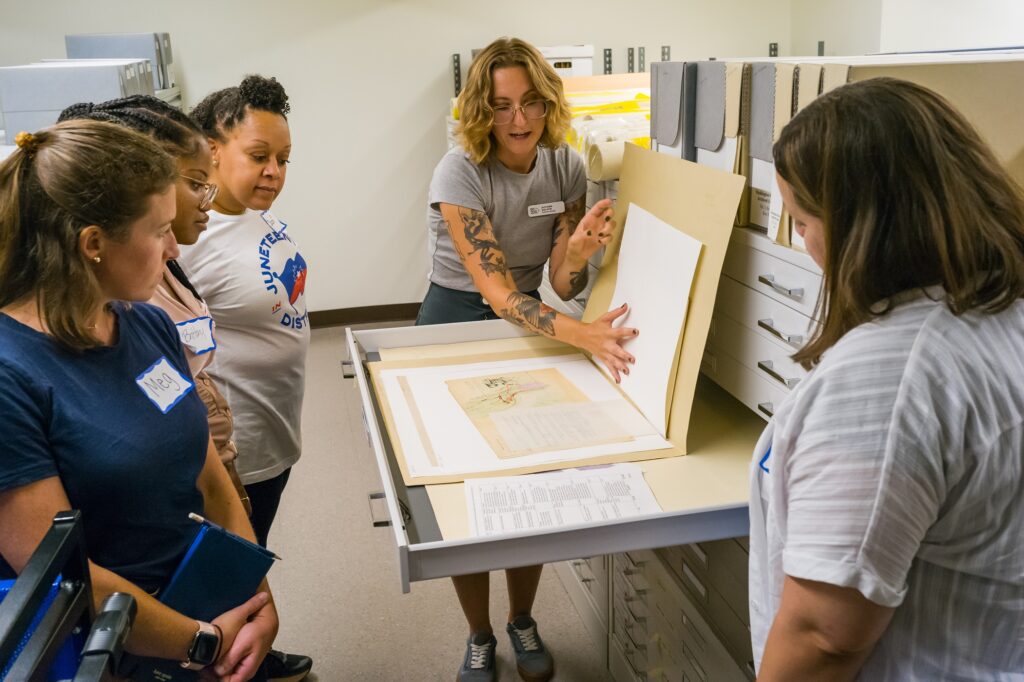 Collections Manager Autumn Kalikin holds up a folder flap in a flat file to reveal a map in the DC History Center archives space. Four female teachers are gathered around and looking at the document.