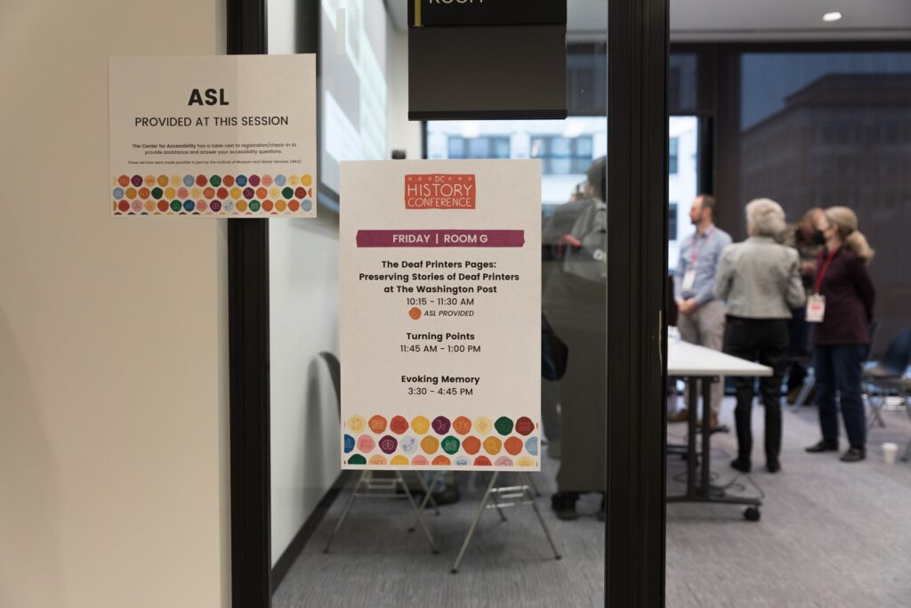 Two paper signs hang beside the door into a DC History Conference session. One reads: ASL Provided At This Session. The other sign lists the room name and the session schedule for the day, including "The Deaf Printer Pages: Preserving the Stories of Deaf Printers at The Washington Post."