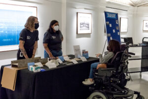 A woman in mechanical wheelchair speaks across a table to two DC History Center staff members during Open Day, a DC History Center event, October 2022.