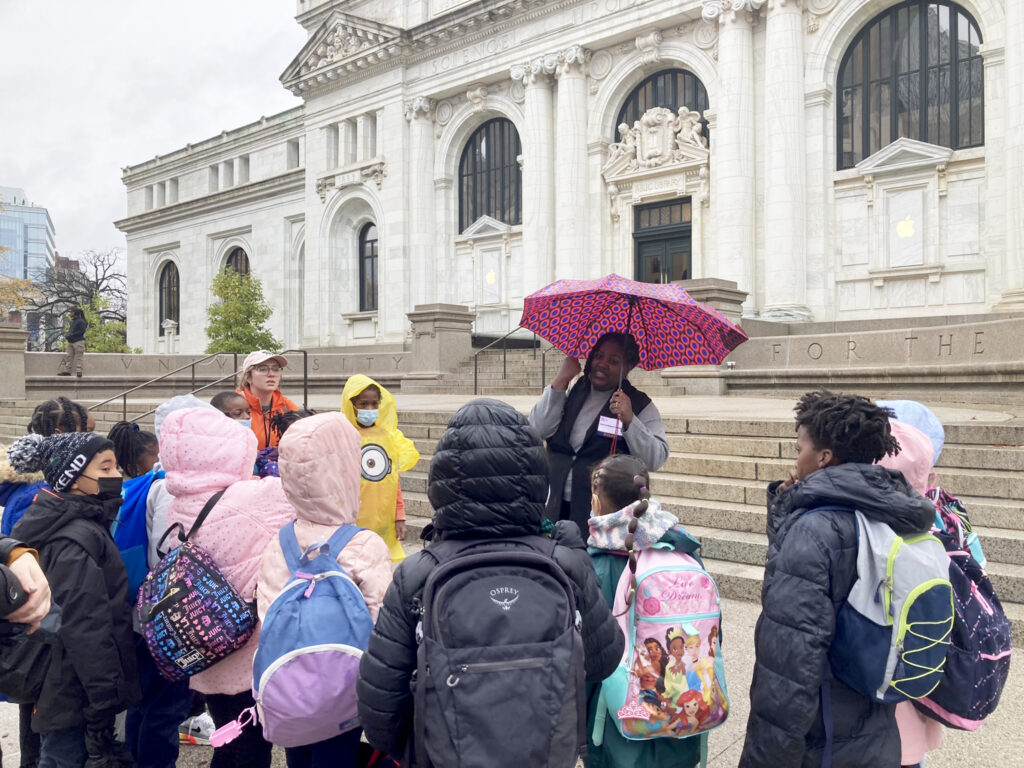 A woman stands under a dark pink umbrella in front of a grand marble building. She is speaking to a large group of third grade students and their teachers wearing winter jackets.