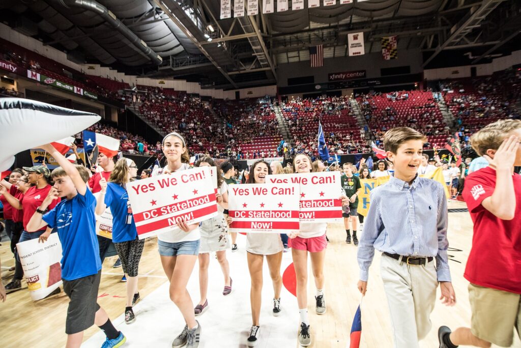 Middle and high school students hold DC Statehood signs that read "Yes We Can! DC Statehood Now!" inside a basketball stadium.