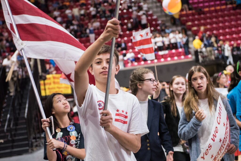 Young male high school student holds up a flag pole inside a large gymnasium. He is wearing a white t-shirt with a small Washington, DC flag printed on the chest. A few female students stand behind him,