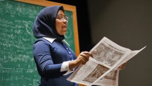 A female highschool student in glassses, historical dress, and a dark blue headscarf reads from a newspaper in front of a chalkboard.