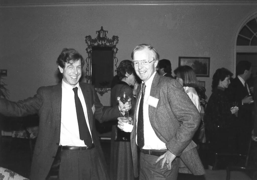 Two men in suits smile and pose with drinks at a social event, surrounded by other guests in conversation. The photo appears to be from the late 1980s, based on clothing and hairstyles.