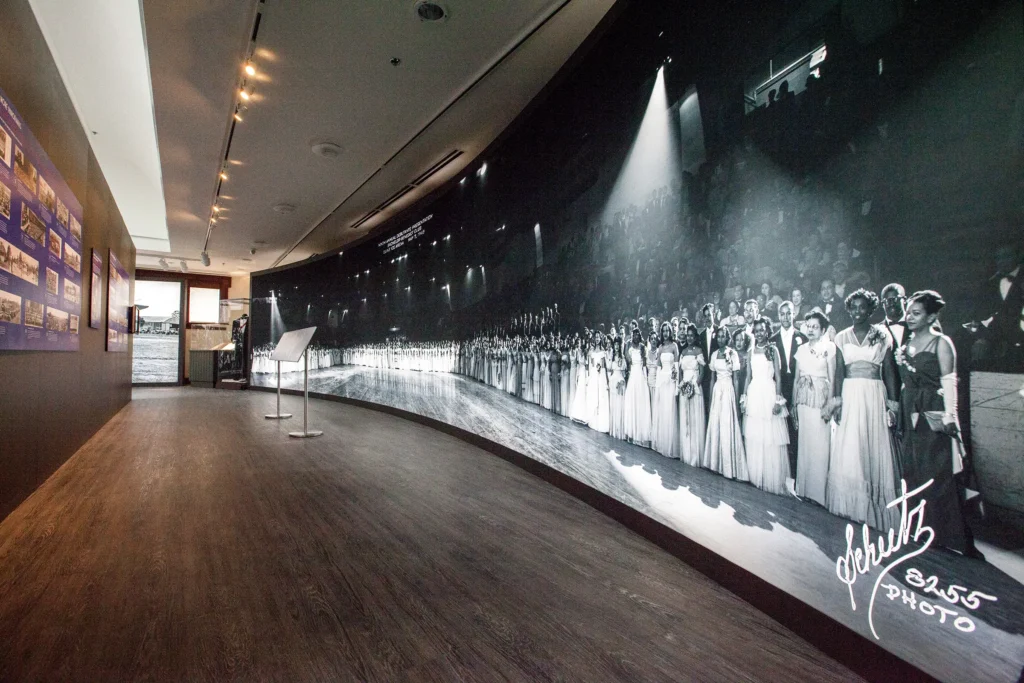 A museum gallery with a curved wall displaying a large black-and-white panoramic photograph of elegantly dressed debutantes and escorts standing in a long line under stage lights. Additional exhibit panels hang on the left wall, and two display stands are positioned in the center of the room.