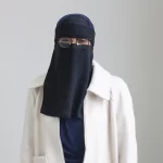 Azzah Gant wears a black veil covering her hair and face, her eyes are visible and she is wearing glasses and a while collared coat.
