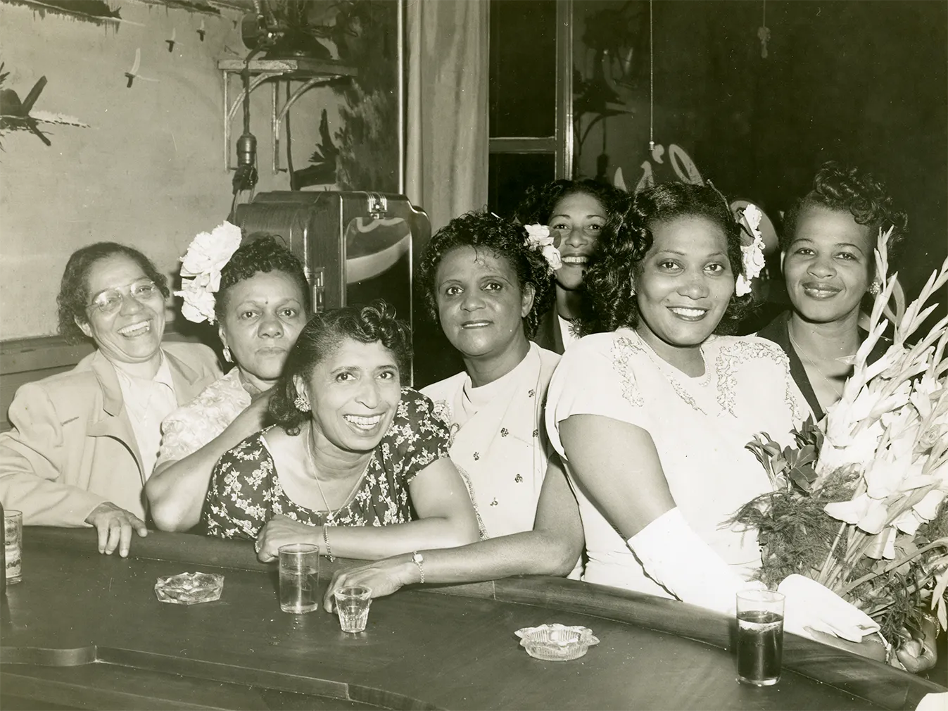 Five women are sat at the bar and two women can be seen behind them, one of the women at the bar is holding a bouquet of flowers. On the bar, three glasses, two ashtrays, and one shot glass are visible.