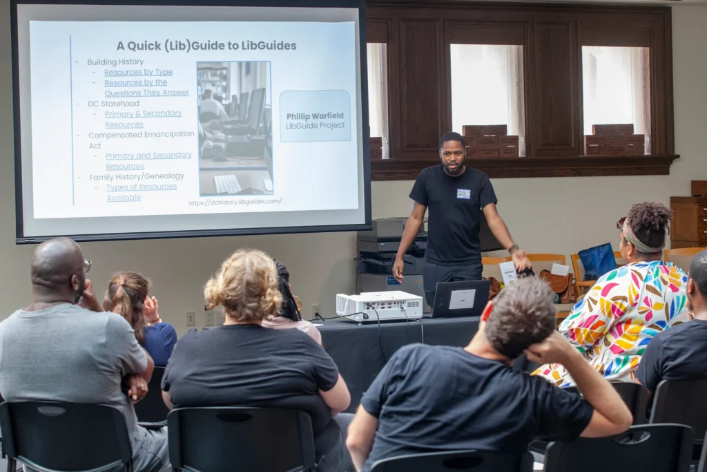 A man with a dark beard and short hair wearing a dark-colored t-shirt addresses a group of seated adults during a presentation. He stands next to a projector screen that reads "A Quick (Lib)Guide to LibGuides."