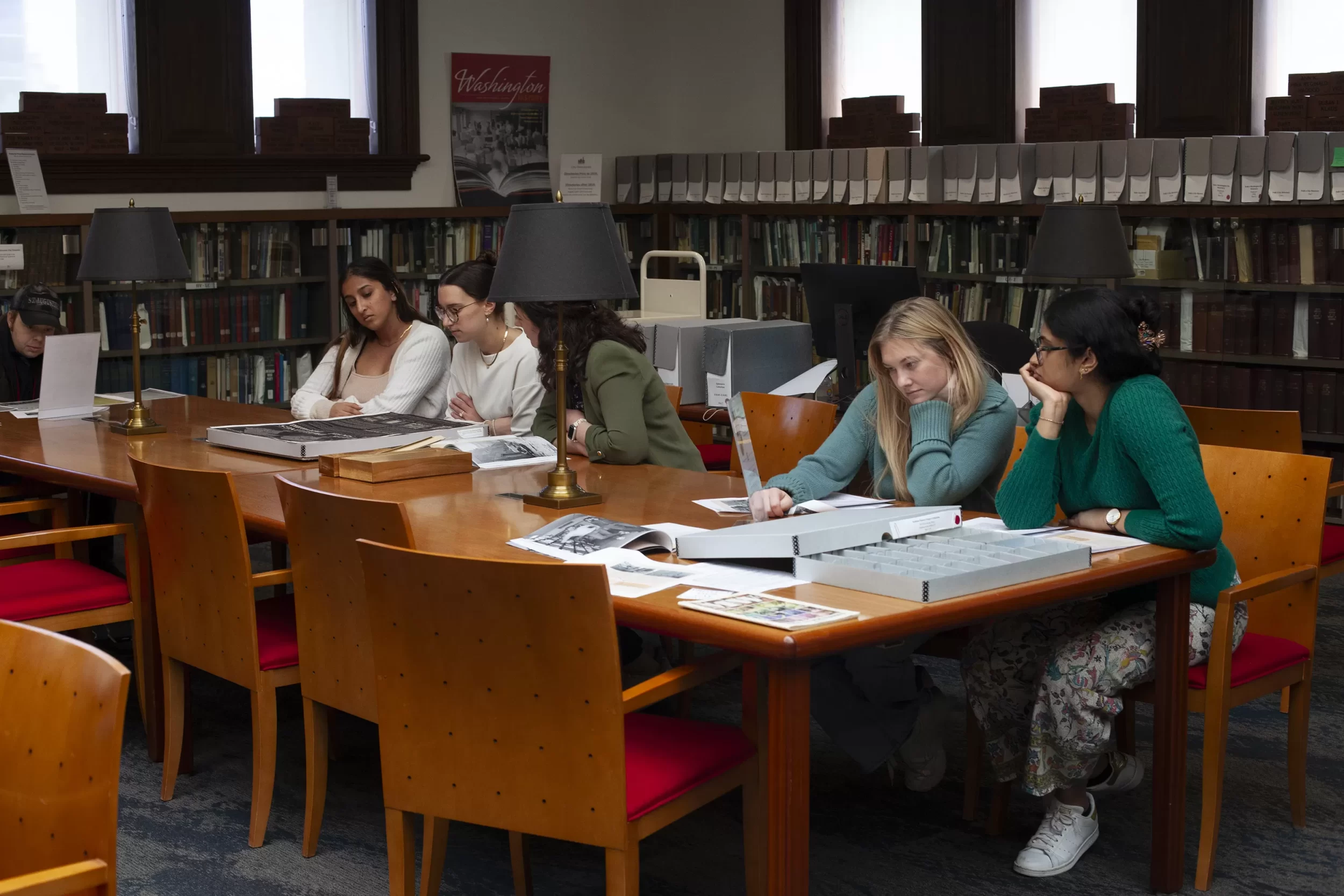 Female graduate students sit at a table in a research library looking at collections material inside gray archival boxes.