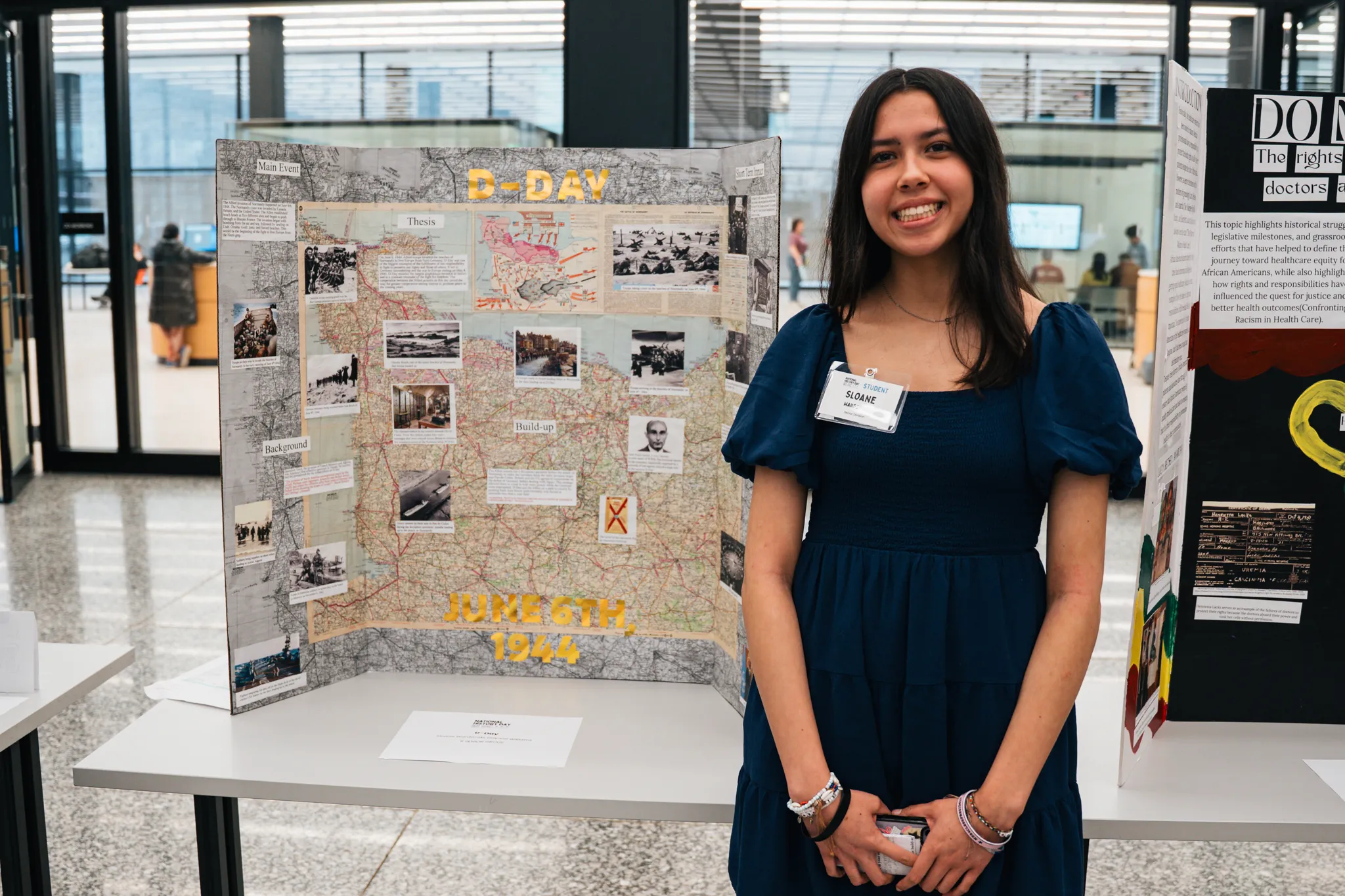 A student stands smiling beside a tri-fold history project display about D-Day, June 6, 1944, featuring maps, photographs, and explanatory text, set up on a table in a modern indoor exhibition space.