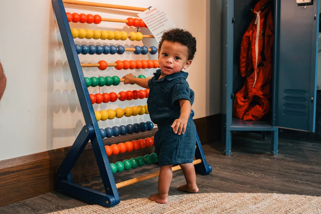 A young toddler stands barefoot on a rug while playing with a large, colorful abacus at the DC History Center’s Class Action exhibit. Behind the child, a vintage school locker with an orange jacket inside adds to the classroom-themed setting.
