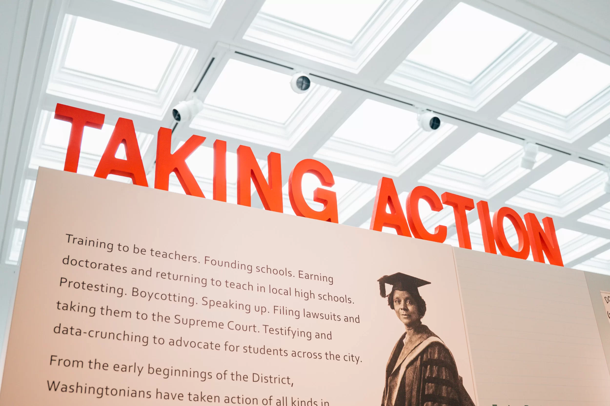 Exhibition panel titled "TAKING ACTION" in bold red letters, with text highlighting educational advocacy efforts and a historic photograph of a Black woman in academic regalia. The display is part of the DC History Center’s "Class Action" exhibition under a skylit ceiling.