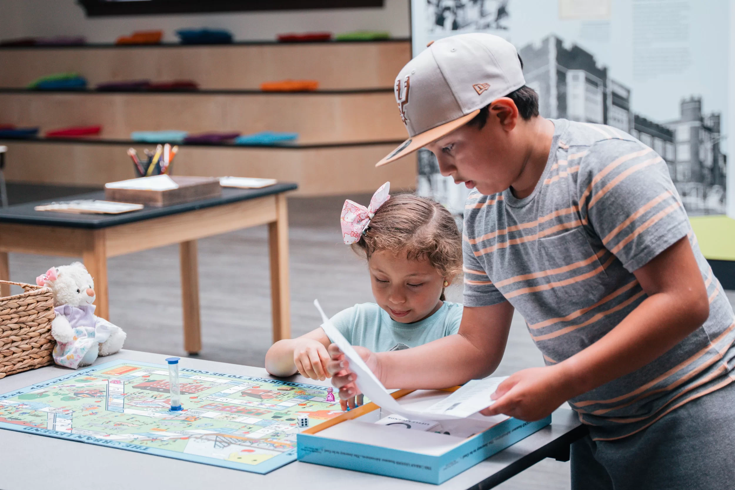 Two young children play a colorful board game together at a table in a museum space. The older child reads instructions while the younger child looks at the board. A plush toy and art supplies are visible nearby, creating a playful and welcoming environment.