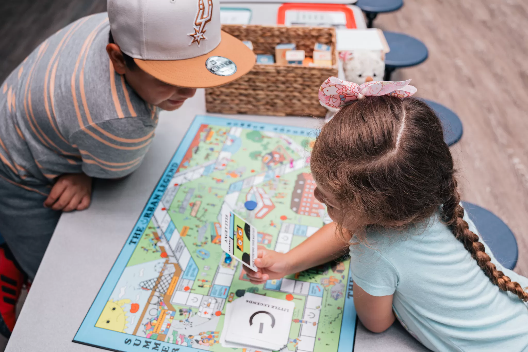 Two young children play a colorful educational board game called "The Urban Legend" at a table. The girl, with a pink bow in her braided hair, holds a card labeled “LITTLE LEGENDS” while the boy in a San Antonio Spurs cap looks on. A woven basket filled with game cards and supplies is visible in the background.