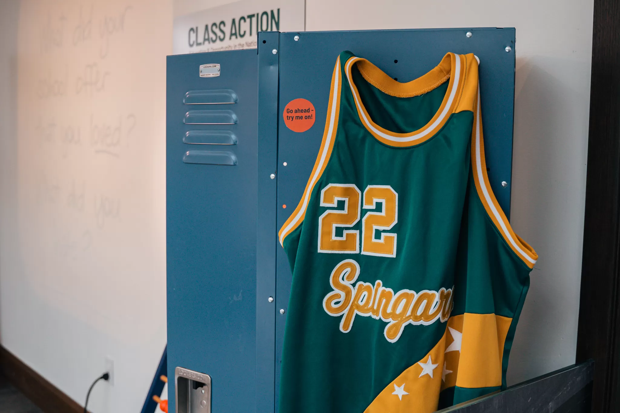 A green and yellow basketball jersey with the number 22 and the word “Spingarn” hangs from an open blue locker in the DC History Center's “Class Action” exhibition. A nearby sign invites visitors to try it on.