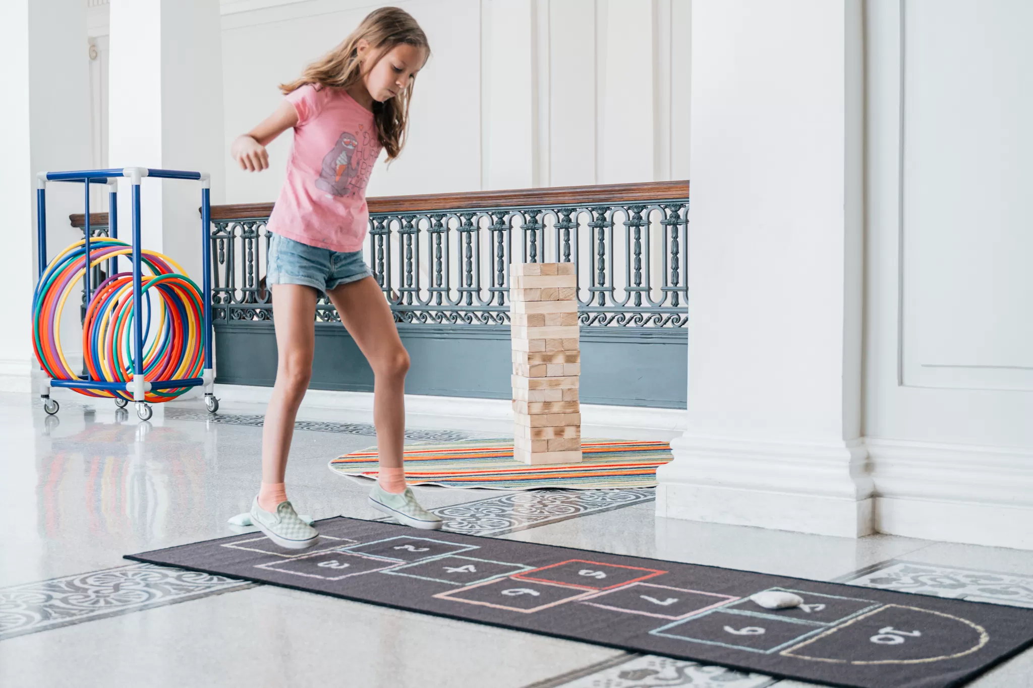 A young girl in a pink tshirt and jean shorts plays hopscotch indoors on a printed rug.