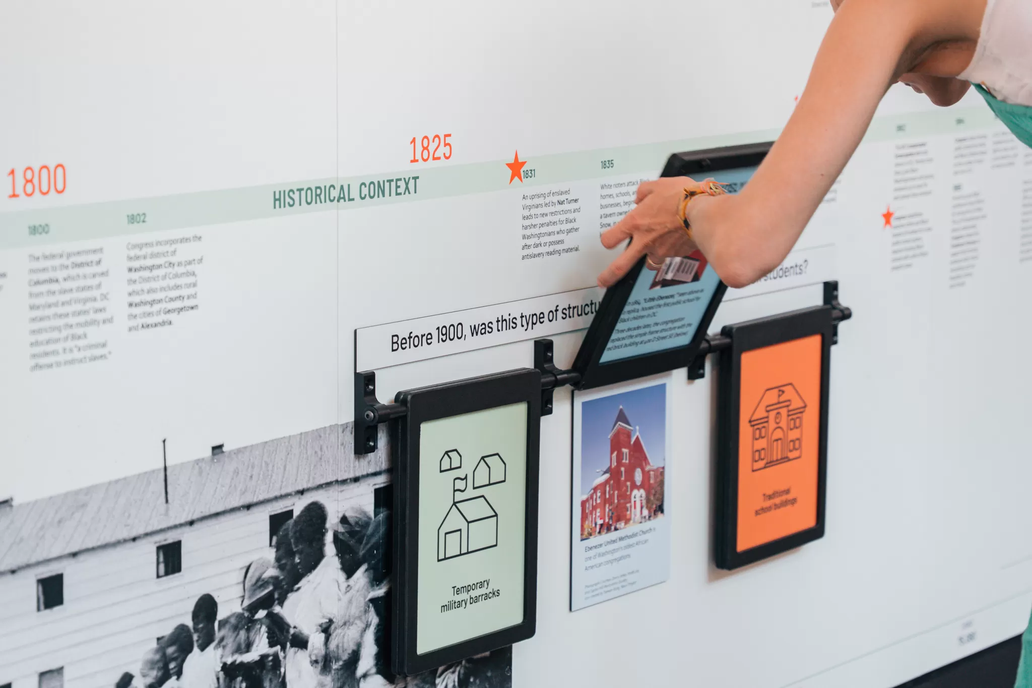 A person lifts a panel on an interactive educational timeline at the DC History Center. The exhibit allows visitors to explore the history of school building types before 1900, with illustrated icons and photos displayed beneath each flap.