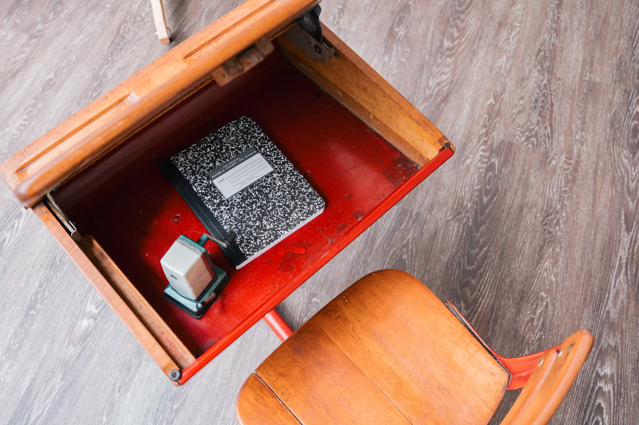 Close-up of an open vintage wooden school desk containing a black-and-white composition notebook and a gray classroom stamp, placed on a wood-patterned floor.