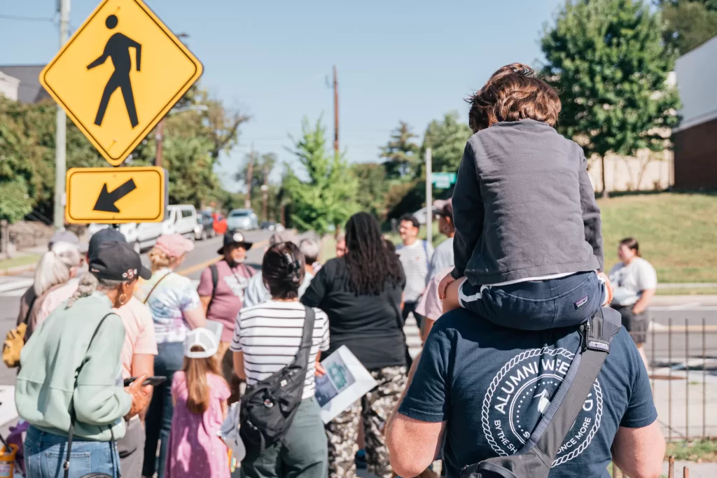 A group of people, including children and adults, gather on a sunny sidewalk near a crosswalk. One adult is carrying a child on their shoulders. A yellow pedestrian crossing sign with an arrow is visible in the foreground. The group appears to be listening to someone speak or lead an activity. The setting is a suburban area with parked cars, trees, and a clear blue sky.