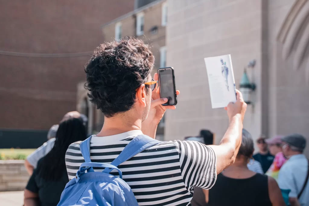 A person with curly hair, wearing a black-and-white striped shirt and a blue backpack, holds up a smartphone and a pamphlet while taking a photo or video. The pamphlet features text and an image of a statue or figure. The scene takes place outdoors in front of a light-colored stone building with architectural details, and a group of people is gathered in the background, some wearing hats and colorful clothing. The mood is sunny and social, possibly part of a walking tour or community event.