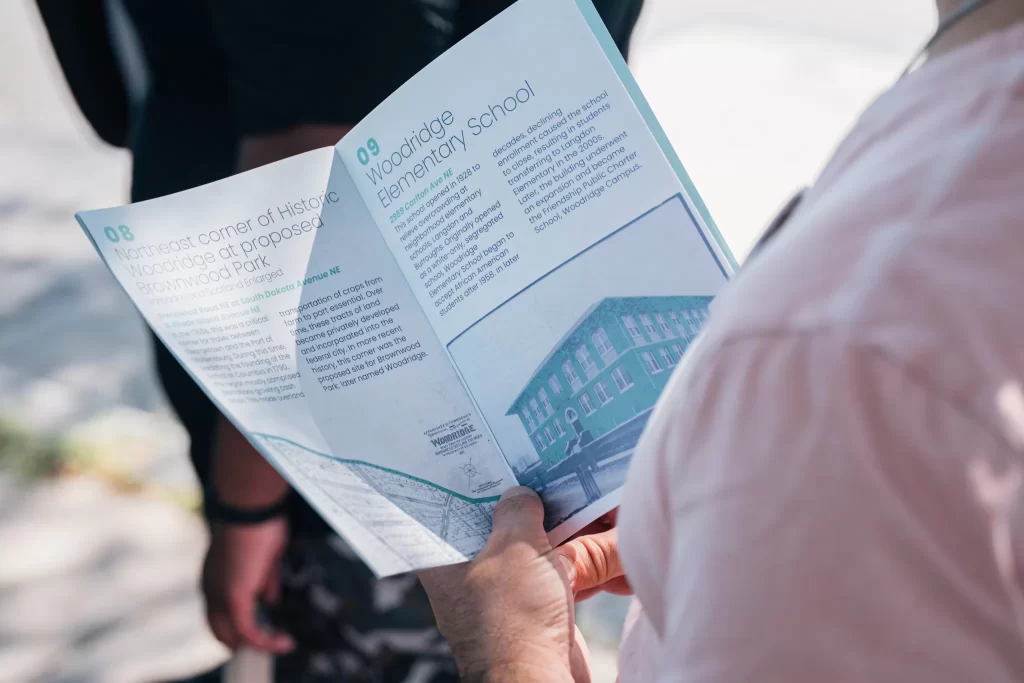 A person in a pink shirt holds and reads a brochure outdoors. The brochure is open to two pages detailing historical landmarks in the Woodridge neighborhood. One page is titled “08 Northeast corner of Historic Woodridge at proposed Brownwood Park,” and the other is titled “09 Woodridge Elementary School.” The pages include historical text, addresses, and a black-and-white photo of a school building. Other people are blurred in the background.