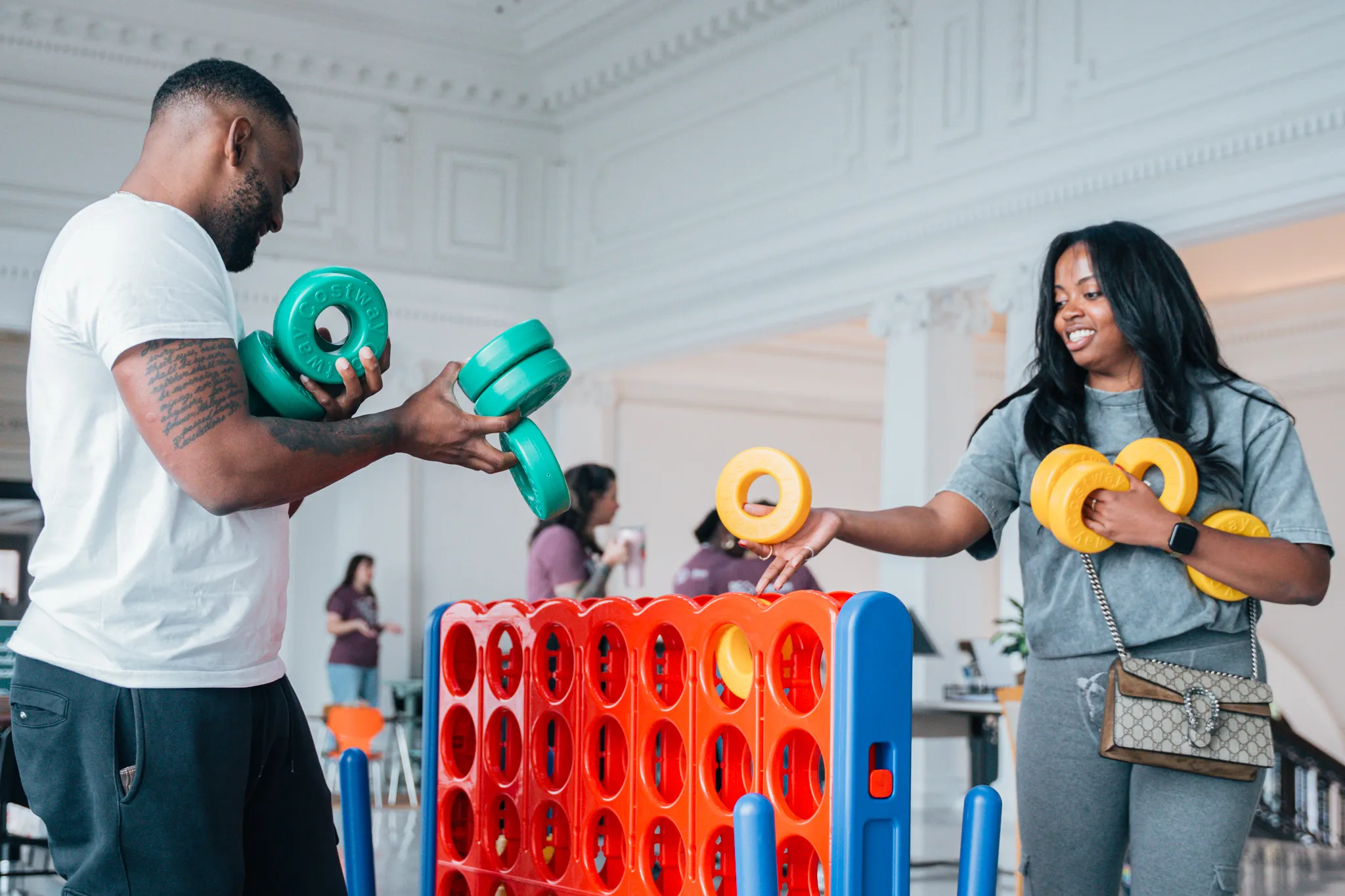 Two adults play a large connect-four game indoors, placing oversized yellow and green rings into a red and blue game board. They face each other and smile, with other people and tables visible in the bright, open room behind them.