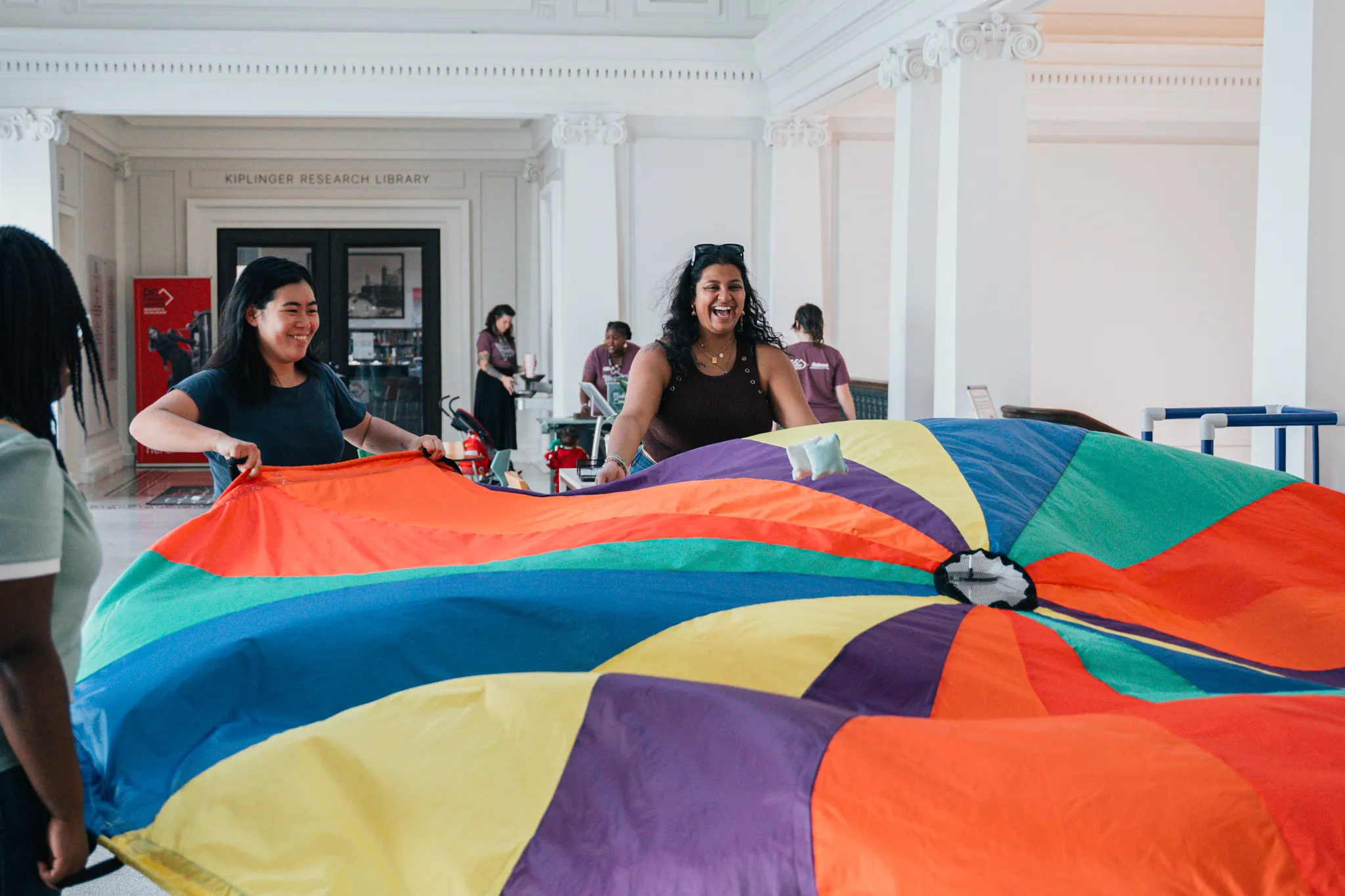 A group of smiling adults play with a large, colorful parachute in a bright, historic hall with white columns. In the background, the entrance to the Kiplinger Research Library is visible, along with other people engaging in activities.