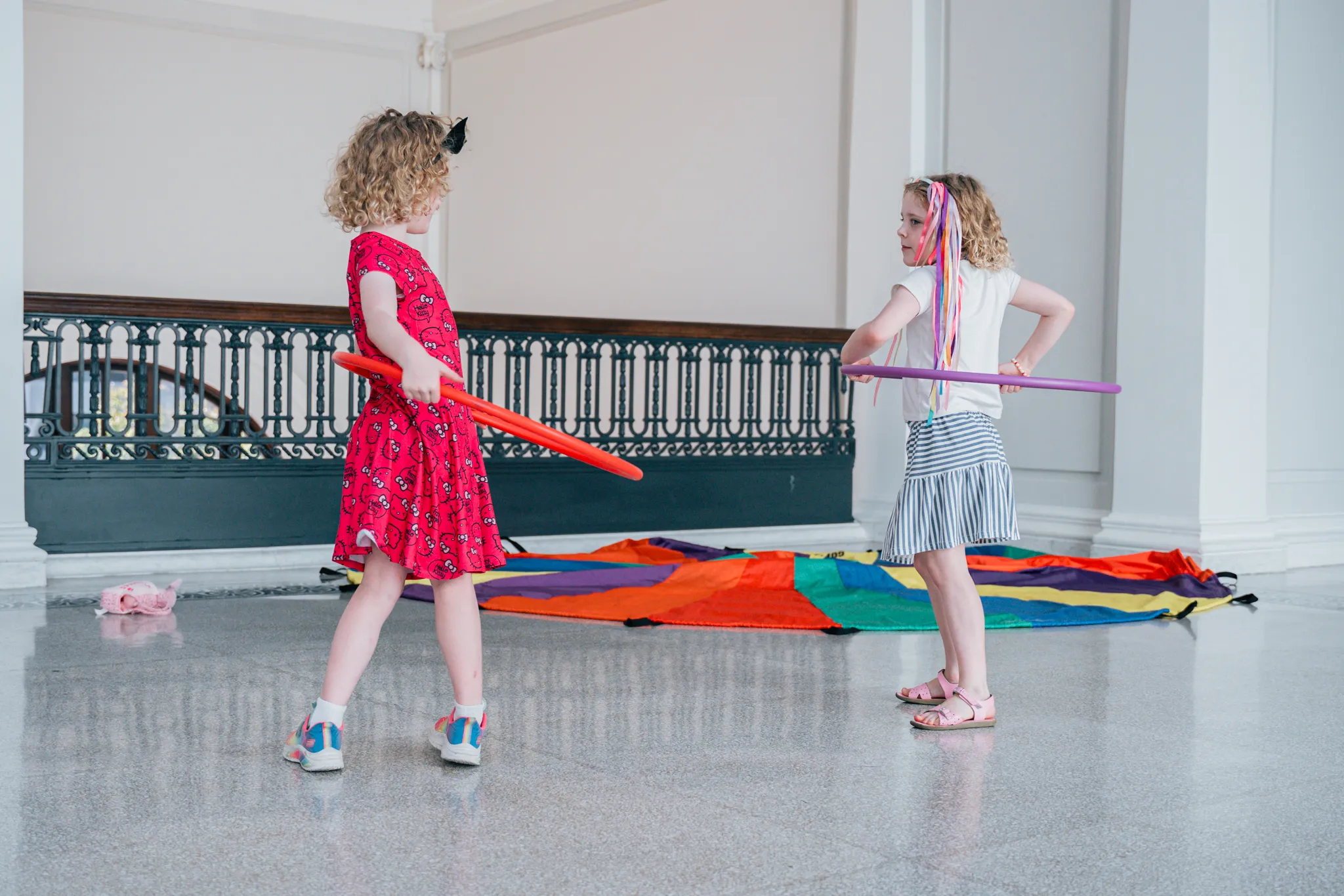 Two young children play indoors with hula hoops, facing each other on a polished floor. One wears a red dress and sneakers, the other a white top and striped skirt with colorful ribbons in their hair. A rainbow parachute lies on the floor behind them near a decorative railing.