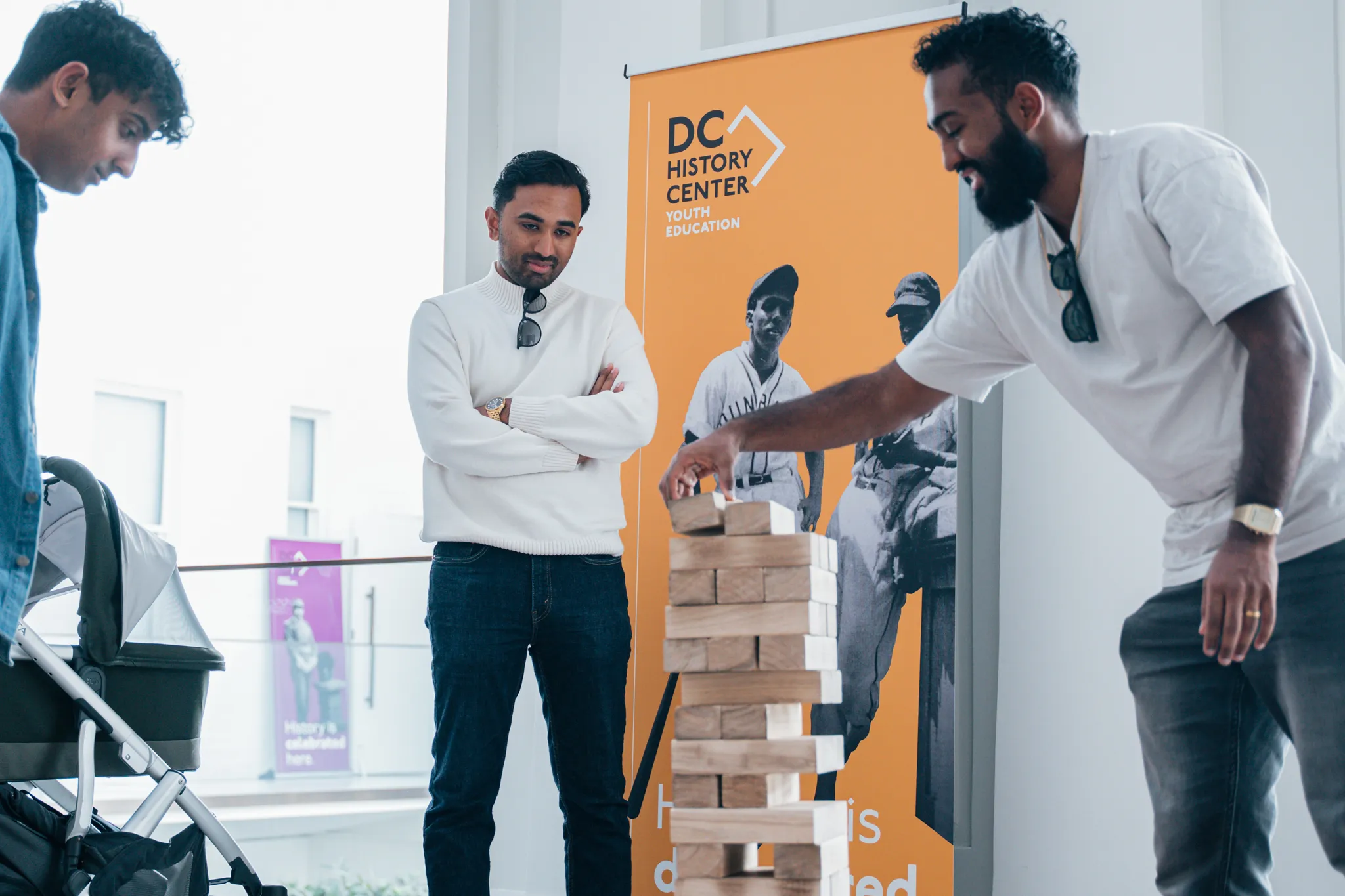 Three adult men play giant sized Jenga in front of a yellow DC History Center pull-up banner.