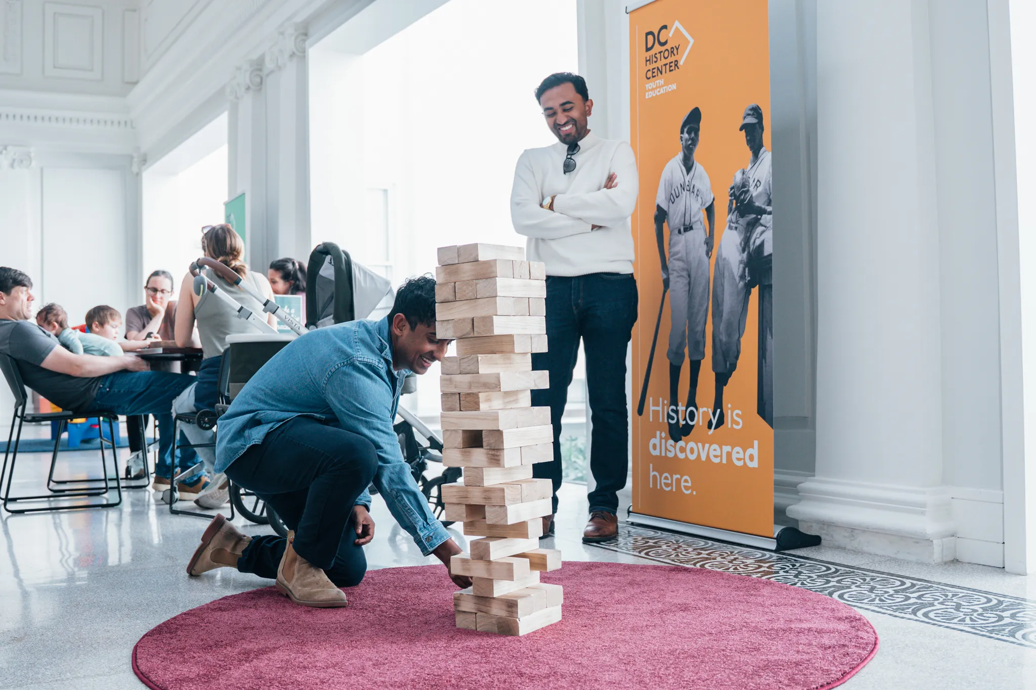 A person kneels on a circular rug indoors, carefully removing a block from a tall wooden stacking game while another person stands nearby watching and smiling. In the background, families sit at tables and a banner reads “DC History Center” with historic baseball imagery.