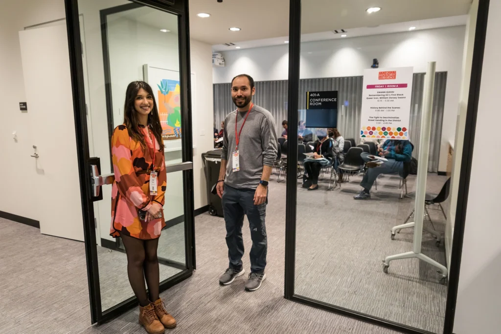 A woman in an orange dress with straight black hair smiles at the camera while holding a glass door open to a conference room. A man with a short dark beard stands in the doorway smiling at the camera, wearing jeans, a gray long-sleeved shirt, and gray sneakers.