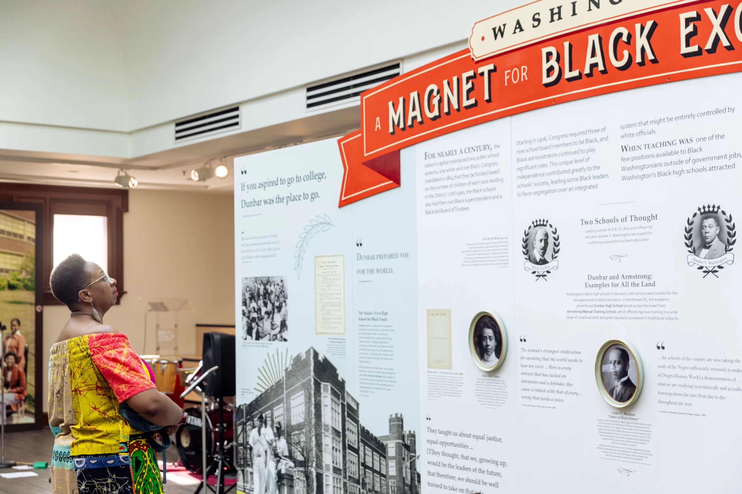 A visitor in a colorful, patterned outfit reads a large exhibition panel titled A Magnet for Black Excellence at the DC History Center. The display features historic photographs, quotes, and portraits highlighting the legacy of Black public high schools in Washington, DC.