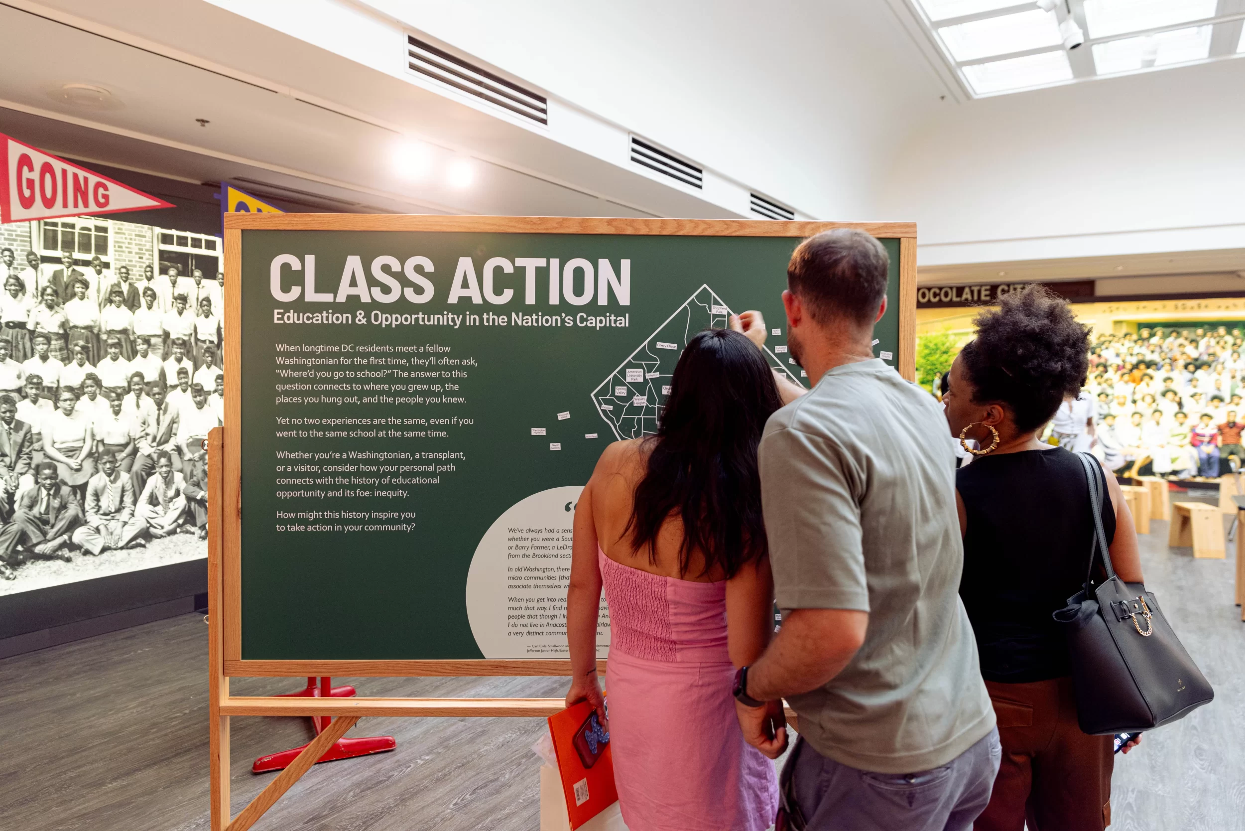 Three visitors—two women and one man—stand closely together, reading and interacting with an exhibit panel titled "Class Action: Education & Opportunity in the Nation’s Capital" at the DC History Center. The green panel includes text and a diagram encouraging personal reflection on educational experiences in DC. Behind them, vintage school photographs and colorful pennants add historical context to the exhibition space.