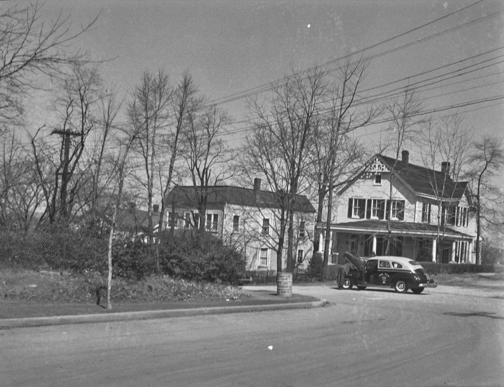Black and white photograph of a quiet street corner featuring two large, old-fashioned houses with visible wooden siding and shutters. Leafless trees suggest it is late fall or winter. A vintage car from the 1930s or 1940s is parked on the roadside with its driver's side door open. Utility poles and wires line the street, and a metal trash can is positioned near the curb. The scene appears to capture a moment from mid-20th century suburban or small-town America.