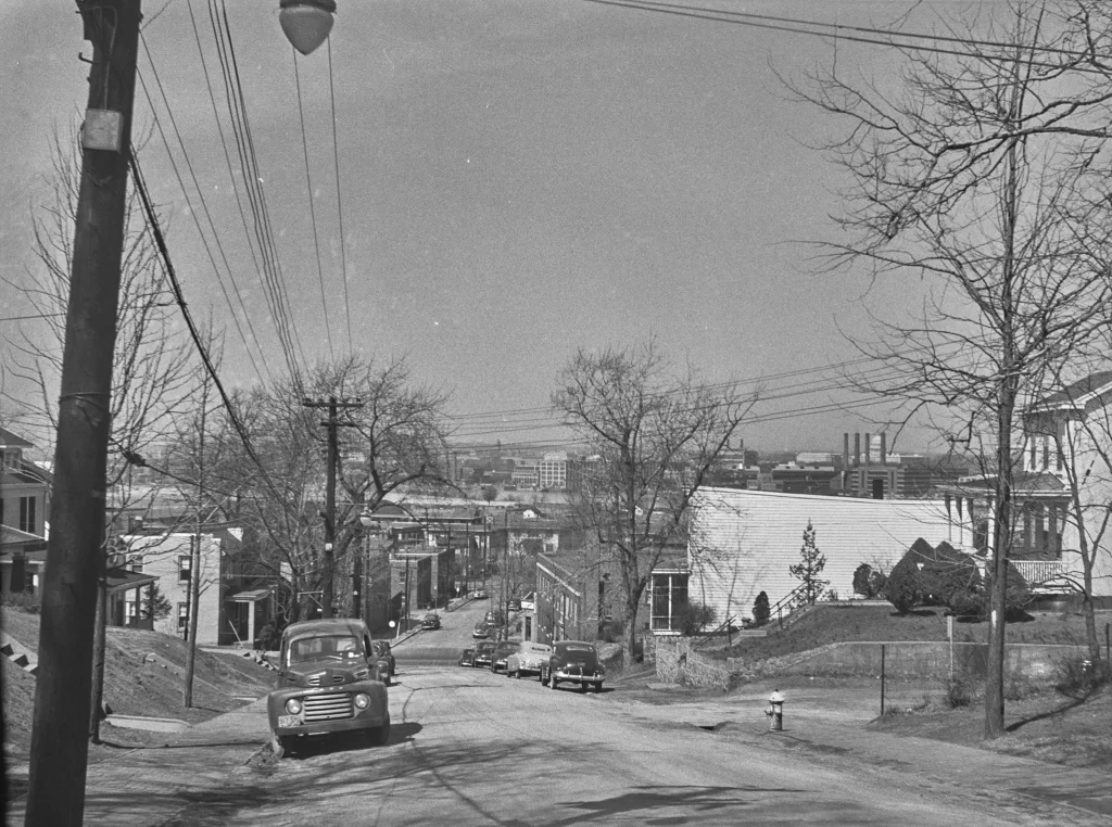 A black-and-white photograph of a residential neighborhood on a hill, likely taken in the mid-20th century. The street is unpaved or lightly paved and lined with utility poles and overhead power lines. Several vintage cars, including a 1940s-era pickup truck, are parked along the street. Leafless trees indicate it is likely late fall or winter. Houses line both sides of the street, and in the distance, an urban skyline with factory smokestacks and mid-rise buildings is visible.
