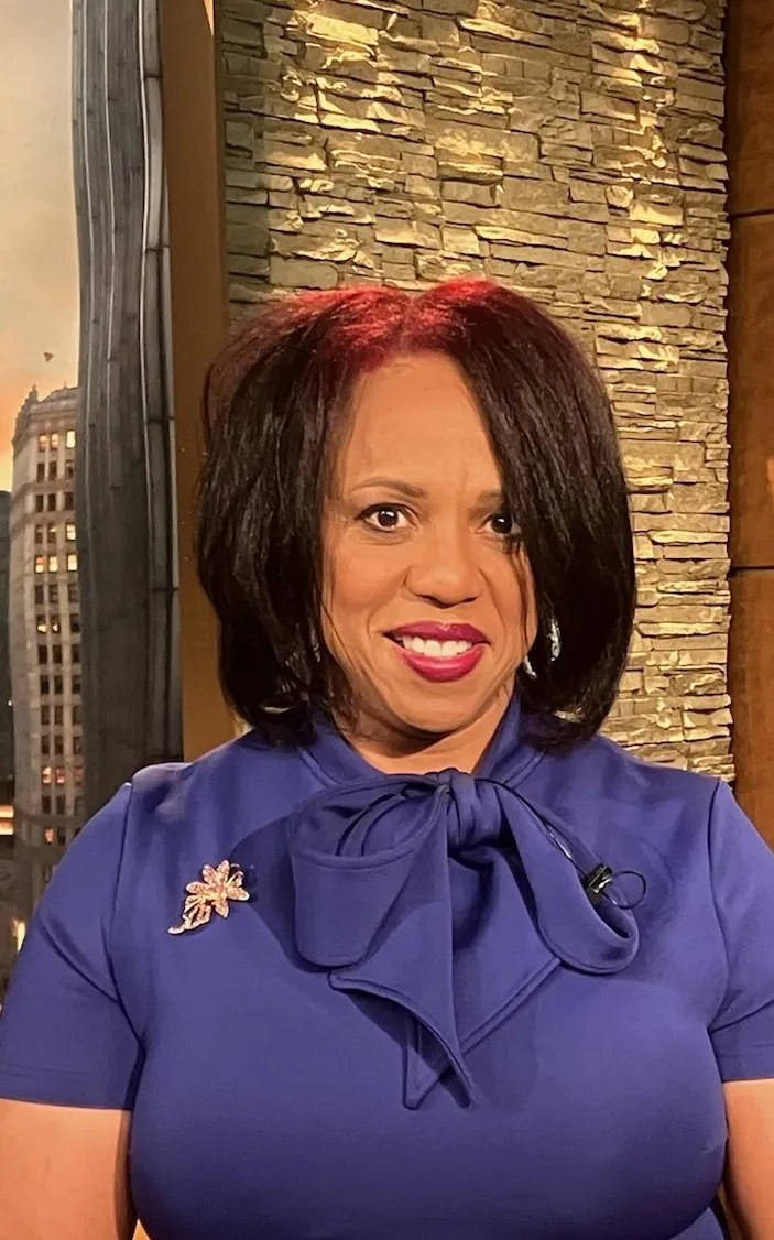 Tikia K. Hamilton, a black woman with shoulder-length dark hair featuring red highlights smiles at the camera in a studio setting. She is wearing a royal blue dress with a bow tie neckline and a decorative brooch on the left side. The background includes a textured stone wall and a softly lit cityscape backdrop, suggesting a television studio environment.