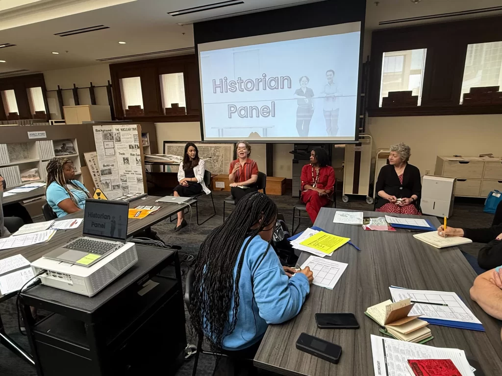 Educators sit around tables listening to a panel of historians speaking in front of a projected slide titled “Historian Panel” during the NHD in DC Summer Intensive.