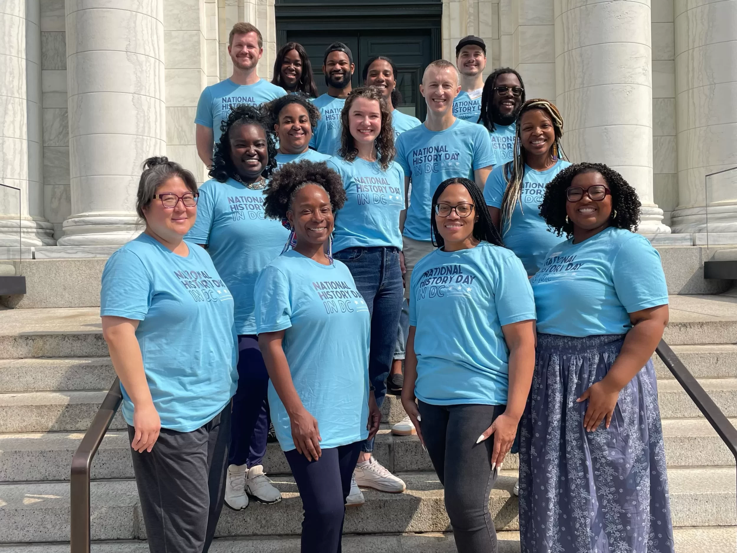 A group of educators wearing matching light blue “National History Day in DC” t-shirts stand on the steps of a marble building, smiling at the camera during the NHD in DC Summer Intensive.