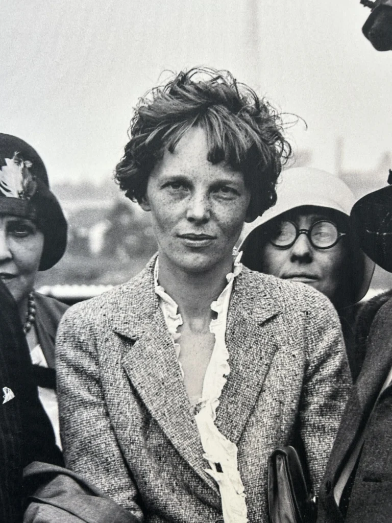 Black-and-white portrait of a woman with short, tousled hair and a freckled face, wearing a textured blazer over a ruffled blouse. She stands outdoors with two other women partially visible behind her.