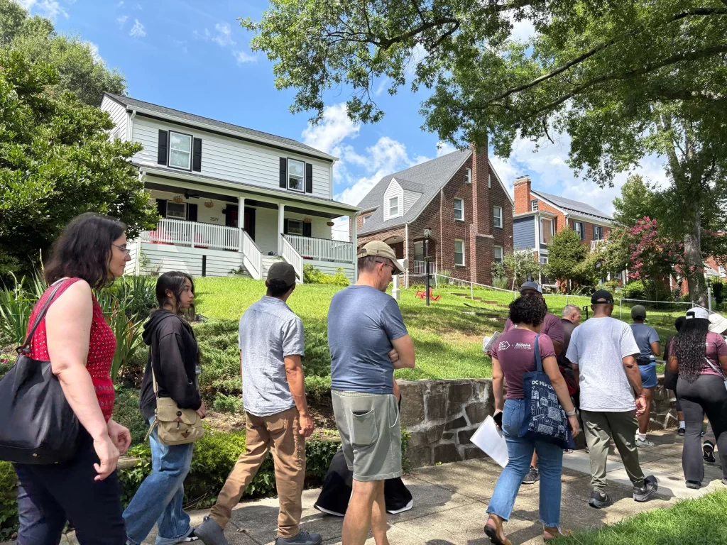 A diverse group of people walks along a sidewalk in a residential neighborhood on a sunny day. They pass by well-kept houses, including a white house with black shutters and a front porch, and several brick homes on a grassy hill. Some people carry papers or wear shirts with logos, suggesting they may be on a guided tour or community walk. The street is lined with trees providing partial shade.