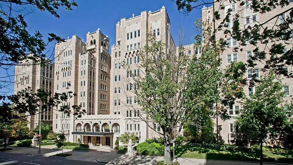 A large historic stone apartment building with multiple towers and arched entrances, seen from across a tree-lined street on a sunny day, with leafy branches framing the foreground and a clear blue sky overhead