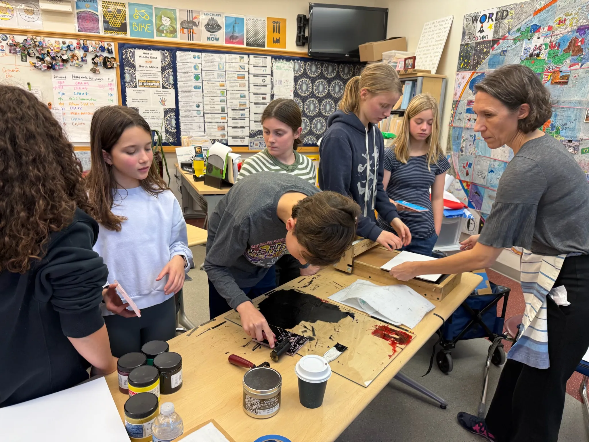 A group of students in a classroom gather around a table while a teacher guides them through a printmaking activity. One student rolls black ink onto a plate with a brayer while others watch and hold printing materials. Ink containers, paper, and tools are spread across the table, and colorful posters, maps, and student artwork cover the classroom walls.