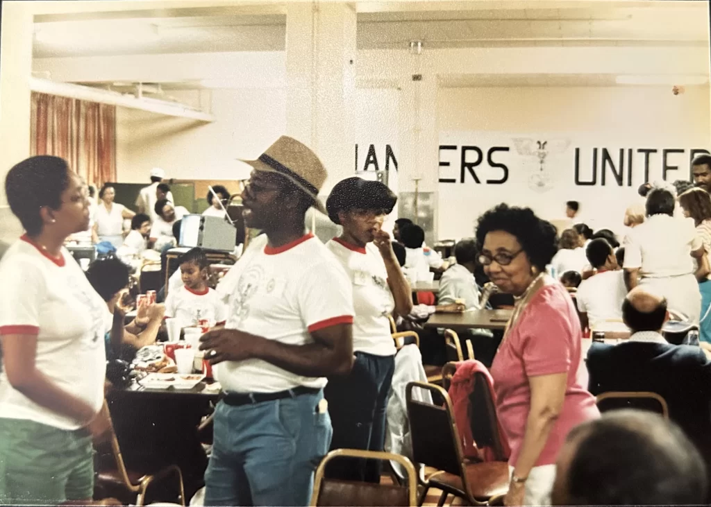 Color photo taken of a large group of African Americans gathered inside a church basement for an indoor picnic, June 1984.