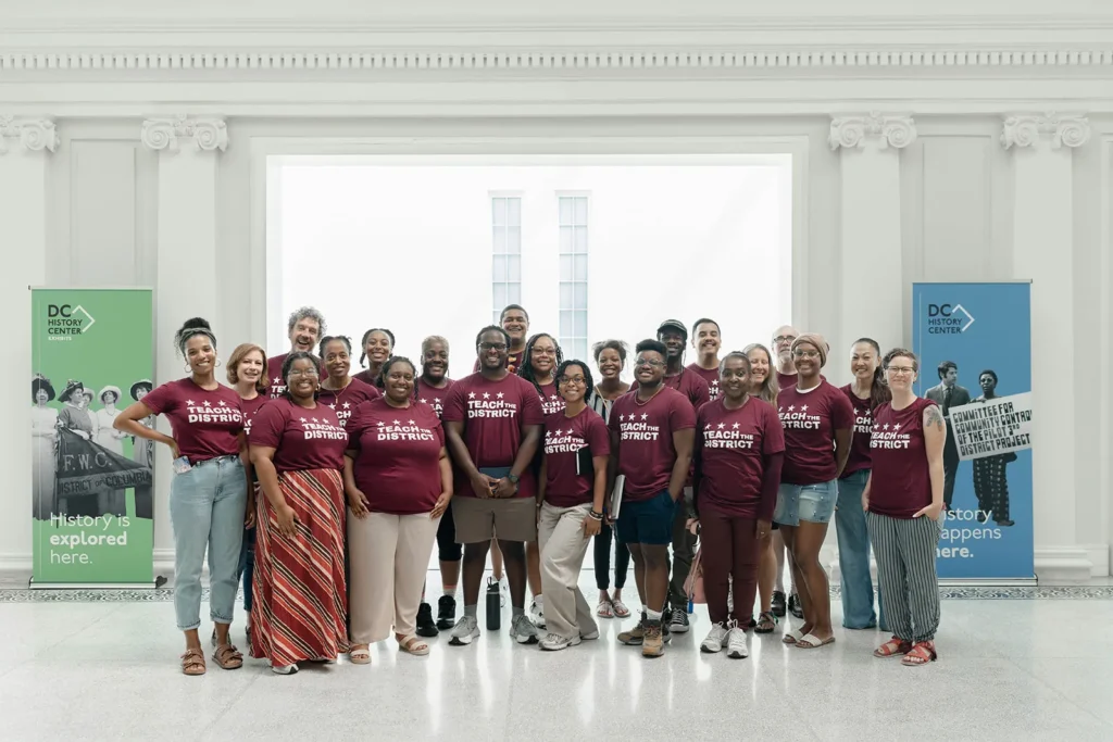 Twenty-one Teach the District educators and DC History Center staff pose together in the DC History Center, wearing burgundy Teach the District T-shirts.